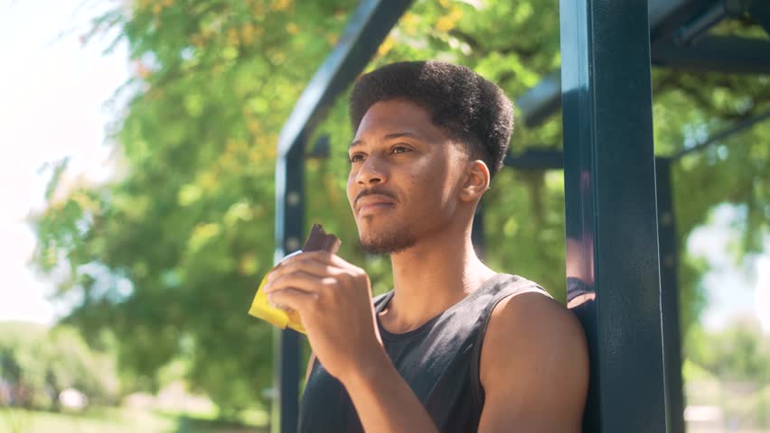 Athletic African American man smiling while eating a chocolate protein bar during a break, captured in a vibrant outdoor park with bright natural light.