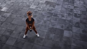 High angle view of focused Black female athlete with curly hair in bun performing kettlebell swing on spacious dark gray tile floor wearing black fitted top with burgundy leggings  - Powered by Shutterstock - Get 15% off with code: PIKWIZARD15