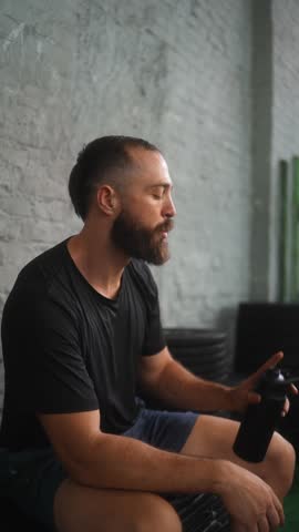 Athletic bearded man in black t-shirt and blue shorts drinking from black insulated tumbler while sitting on exercise equipment in functional gym with white brick wall