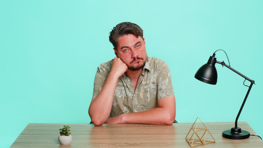 Middle-aged man sits at table with blank stare, head supported by hand, showing emotional burnout, tiredness, complete apathy. Young adult guy isolated on blue background expressing exhaustion fatigue