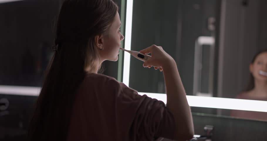 Teenage girl brushing her teeth and rinsing her mouth during her morning routine in front of a mirror in a modern bathroom. Monitoring the cleanliness of teeth