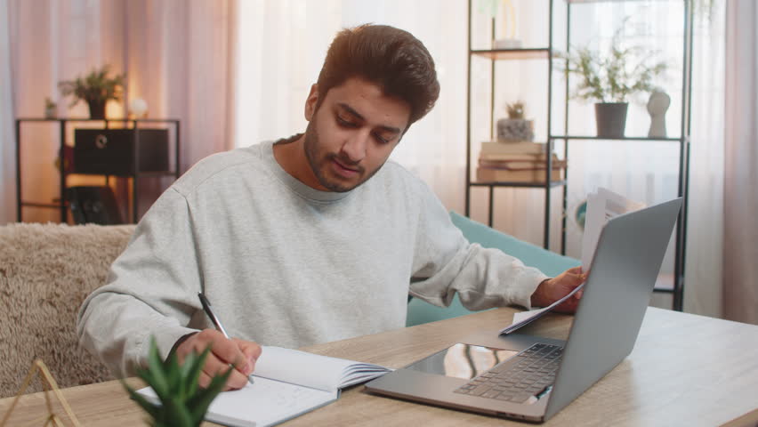 Young Indian man freelancer sitting at table reviewing financial reports on laptop, holding documents and receipts, taking notes in notebook. Hispanic guy sits nearby at home, focused on his own task.