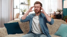 Young man at home sits on sofa, holding his head with hand and massaging his temples in discomfort. Caucasian guy suffers from a strong headache, feeling stressed, tired, or overwhelmed, bad day - Powered by Shutterstock - Get 15% off with code: PIKWIZARD15