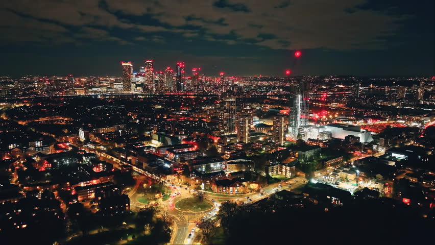 Breathtaking aerial view of London at night, highlighting the vibrant glow of Canary Wharf amidst a bustling urban landscape filled with illuminated buildings and dynamic energy. Drone flight footage