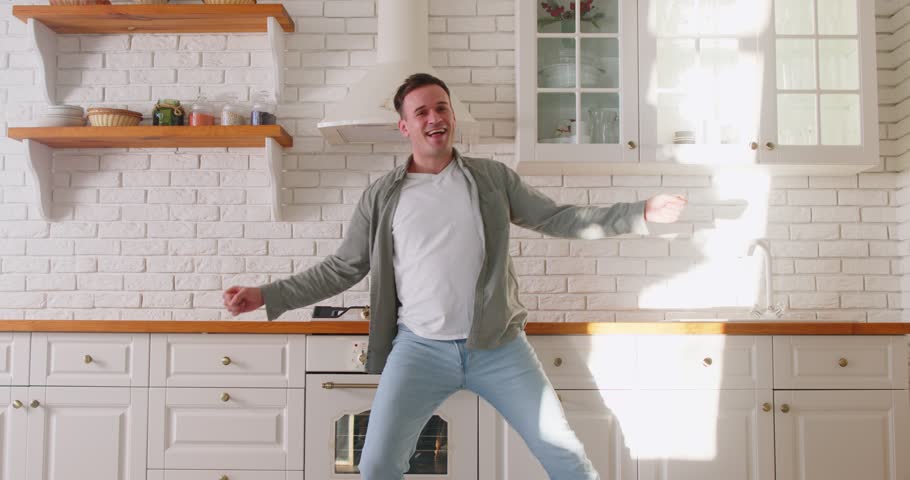 Young happy excited overjoyed man wearing casual clothes dancing in modern kitchen at home. Guy having fun with a satisfied face expression enjoying good mood. People emotions concept. 4k video.