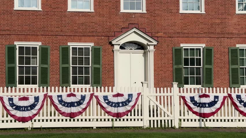 Establishing shot of a small-town red brick building with colonial architecture. Green shutters, white picket fence, and patriotic bunting evoke classic Americana charm in a quaint village setting. 14