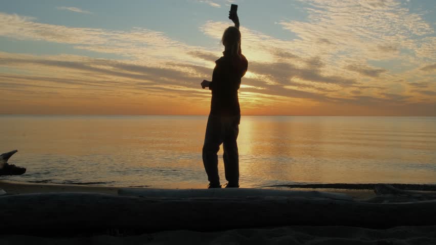 Silhouette of woman searching for mobile phone signal or gps coverage on her smartphone while walking on the seashore at sunset.