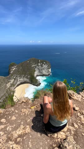 Woman enjoying breathtaking view of Kelingking Beach and turquoise ocean from cliff viewpoint on Nusa Penida, Bali. Perfect for travel, freedom, summer, and adventure themes.
