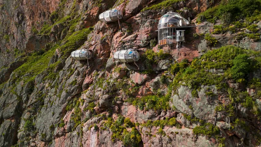An aerial tilt-up shot reveals the famous Skylodge Adventure Suites, transparent glass capsule pods hanging precariously from a sheer cliff face in the Sacred Valley of Peru, an extreme hotel.