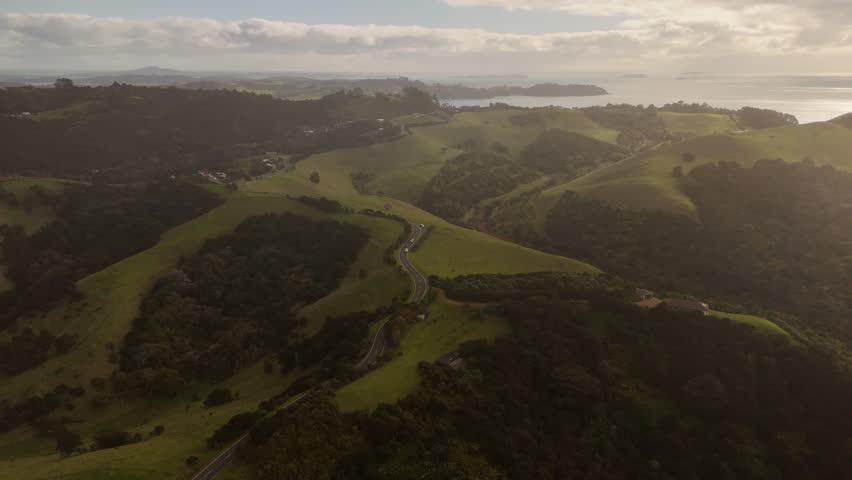 Cars on scenic road winding through lush green hills of Stony Batter Reserve, Waiheke Island coastline is visible, New Zealand. Aerial forward
