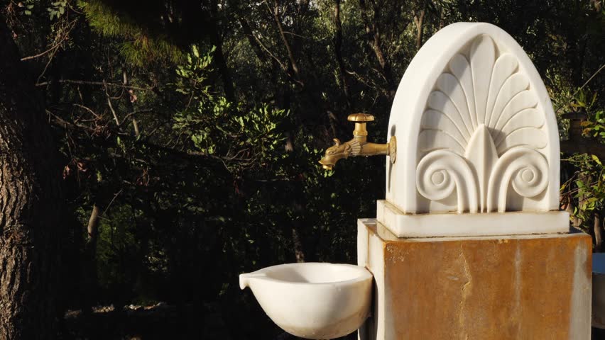 Detailed close-up of a traditional Greek marble water fountain near the Odeon of Herodes Atticus in Athens. Shows ornate brass spout, carved shell, and floral designs. No people visible.