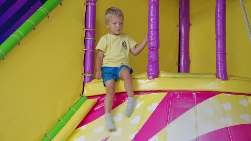 A young boy is sitting and holding onto a purple padded pole. He is inside a colorful indoor playground filled with soft structures and vibrant colors.