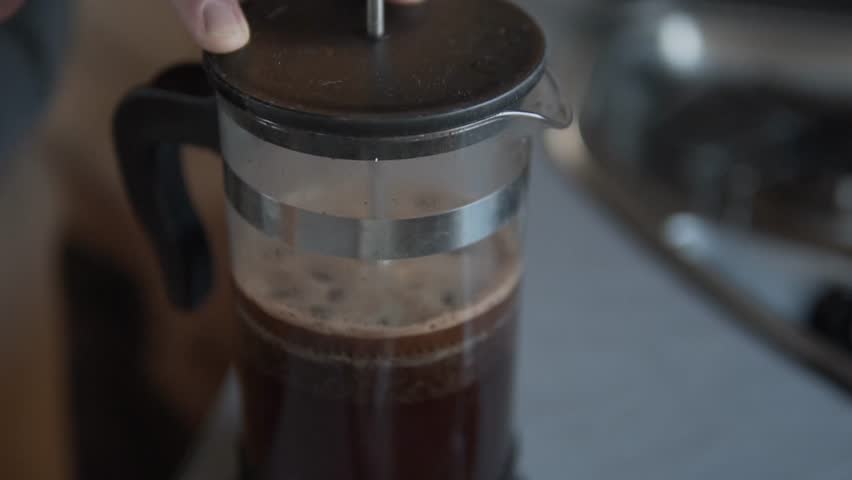 A close-up view of hands gently pressing the plunger of a clear glass French press, brewing rich, dark coffee with visible grounds in a cozy indoor setting.