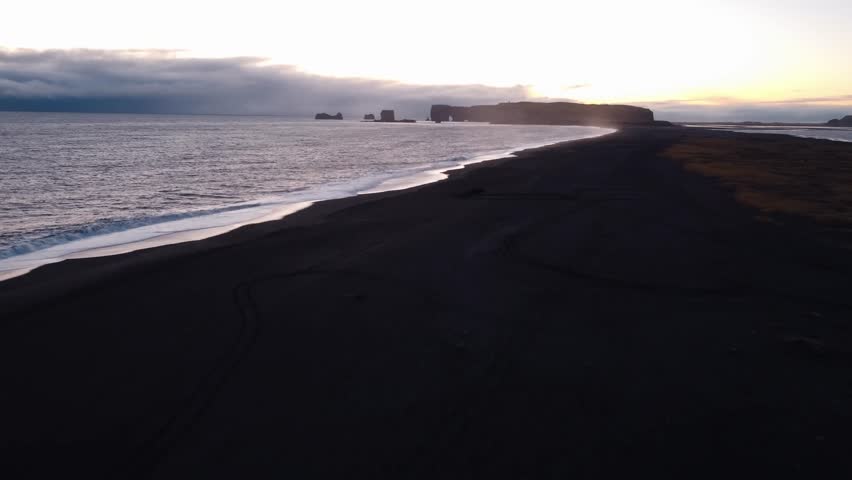 A tranquil black sand beach at Reynisfjara, Iceland, stretches into the distance, reflecting the soft hues of a sunset or sunrise over the calm North Atlantic waters.