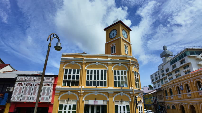 Timelapse Daylight  A tranquil daytime view of Phuket’s colonial landmarks, with crisp white clouds scattered across the deep blue sky. The clock tower and buildings radiate character and charm