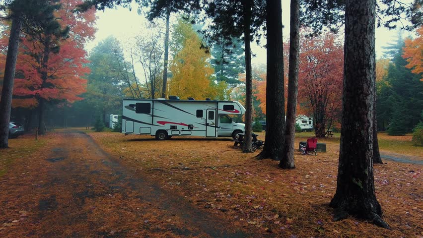 Adirondack Mountain, New York State. An RV parked in a wooded area during autumn. The trees have a mix of red, orange, and yellow leaves, and the ground is covered in fallen leaves.
