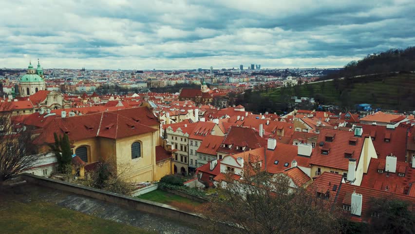 Prague. Aerial view of historic European cityscape with redroofed buildings and green rooftops, set against a partly cloudy sky.