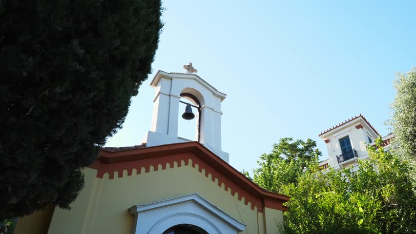 Part of a church with light walls, orange-brown frieze, and decorative pattern. Bell tower topped with cross. Surrounded by dark wood and green foliage, with a clear blue sky above. No people