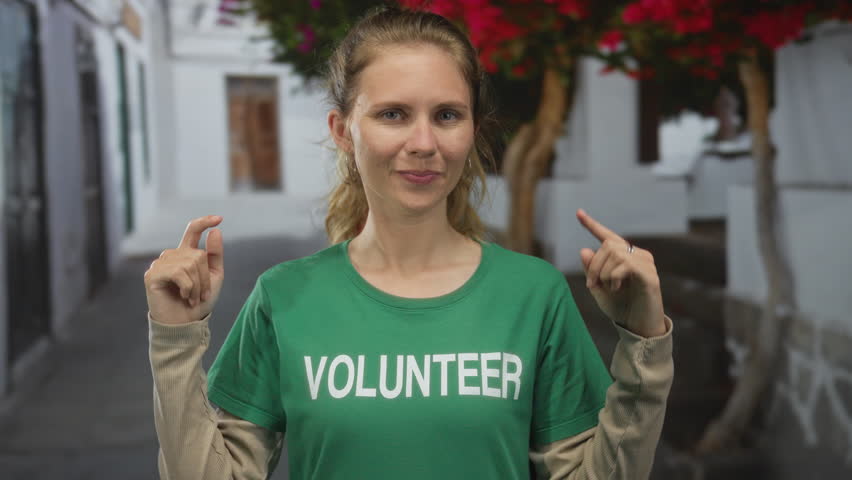 Woman holds out hands pointing towards a white street while a blonde young volunteer in a green shirt shows a warm smile and brushes hair behind ear.
