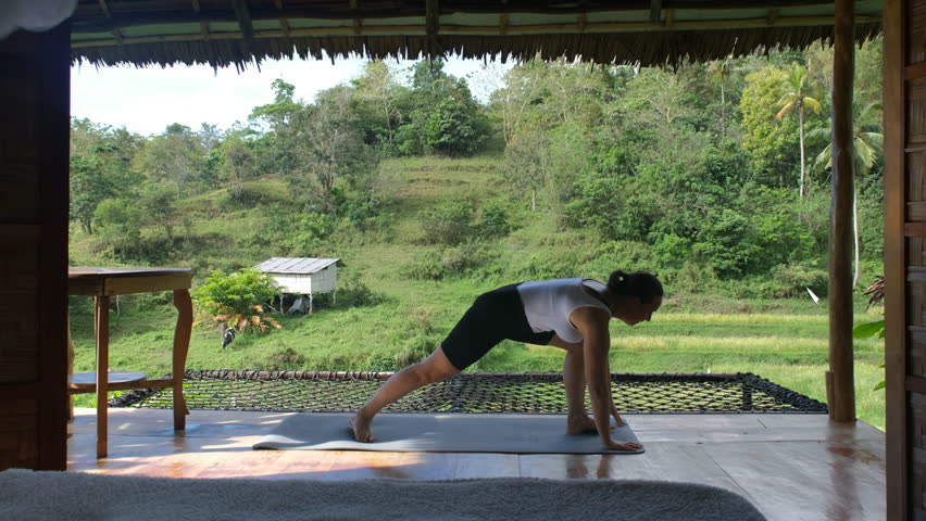 In a tranquil environment, a young woman engages in yoga on a bamboo terrace, surrounded by vibrant tropical greenery and rice plantations, wellness and active lifestyle. Slow motion