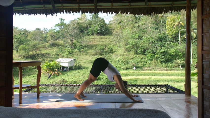 In a tranquil environment, a young woman engages in yoga on a bamboo terrace, surrounded by vibrant tropical greenery and rice plantations, wellness and active lifestyle. Slow motion
