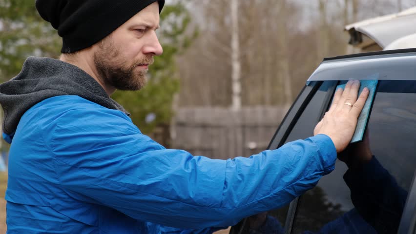 Guy cleaning car in his backyard. Handsome bearded man washing car by hand, cleaning the car interior. Close up of mid adult man hand with a polish cloth, a sponge. Manual vehicle wash outside.