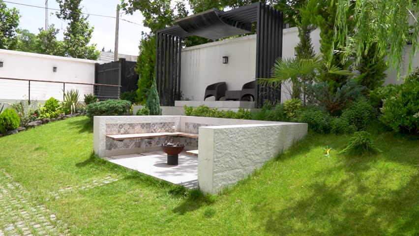 Modern landscaped garden with tiered grass levels, built-in concrete seating, and a central fire pit or table area. Surrounded by lush green plants, black fencing, and trees, this backyard space