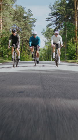 Group of strong cyclists wearing helmets rides along country road across wood. Men athletes race on professional bicycles through summer pine forest