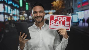 Young man holding a tablet and a for sale sign smiling on an urban street at night reflecting real estate technology in the cityscape - Powered by Shutterstock - Get 15% off with code: PIKWIZARD15