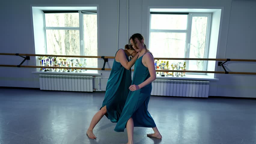 Couple of pretty Caucasian women in similar blue dresses perform a contemporary ballet dance in a bright studio at daytime. Ladies are performing mix of choreography and freestyle improvisation