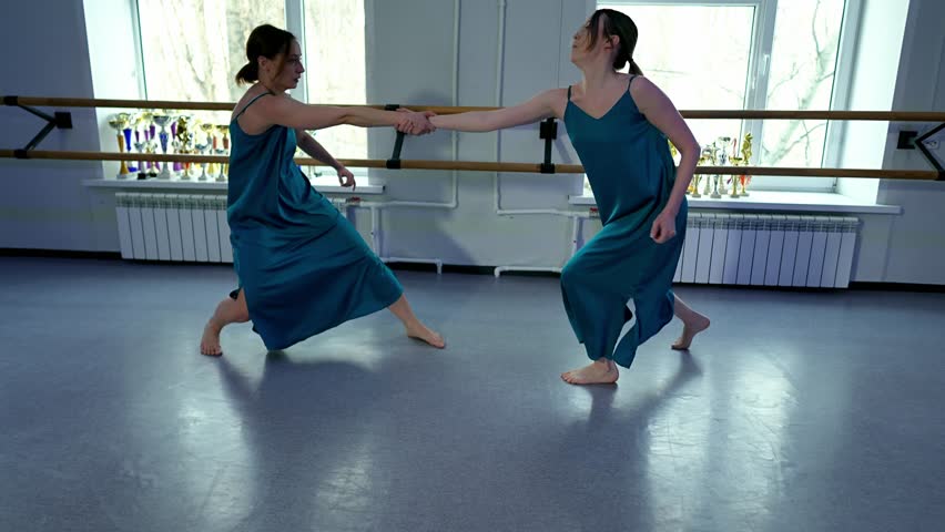 Two young adult Caucasian women dance barefoot in a studio showcasing a modern ballet performance. Wearing blue dresses they are exercising in artistic expression through freestyle choreography