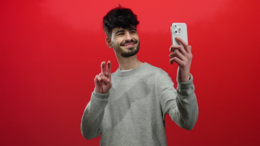 Young man taking selfie over red background using smartphone smiling confidently while showcasing peace sign and wearing casual gray sweater
