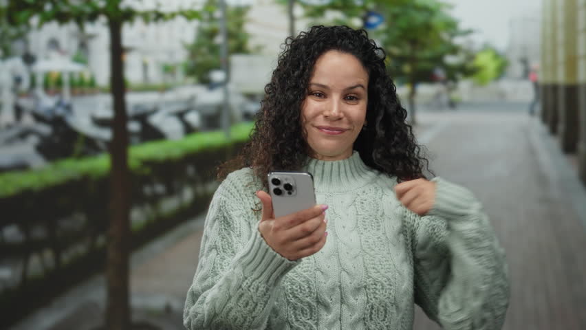 Young hispanic woman smiling while pointing to herself and holding a smartphone on an outdoor street setting, wearing a knit sweater against a blurred urban background.