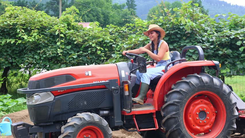 Young Latin female farmer driving red tractor across agricultural field, wearing straw hat, bib overalls, and work boots with green landscape surrounding her