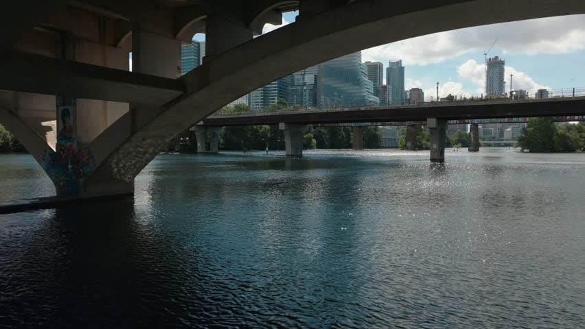 Drone flies out from beneath Lamar Bridge and rises over Ladybird Lake, revealing the Austin skyline and pedestrian bridge in golden light on a beautiful summer morning