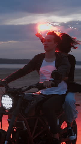 Couple on vintage motorcycle with red burning signal fire riding after sunset on beach, slow motion