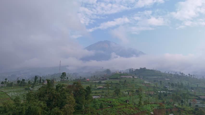 Scenic aerial shot of terraced fields and rural buildings surrounded by thick fog and low-lying clouds over the mountains. Tropical rural landscape, Indonesia.