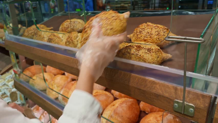 selecting freshly baked bread from bakery display case. baker's gloved hands carefully place a freshly baked, crusty loaf of bread, sprinkled with seeds, into a warm display case filled artisan bread