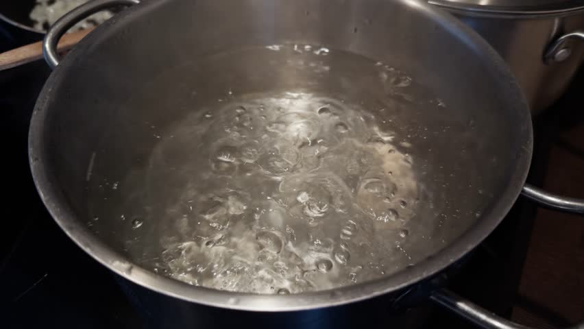 boiling water in a stainless steel pot on a stovetop, with bubbling motion captured mid-cook, and chopped onions frying in a pan in the background, representing food preparation