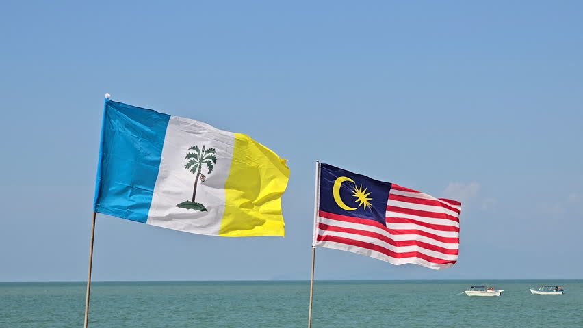 A scenic coastal view featuring the Penang state flag and Malaysian national flag fluttering in the sea breeze with boats in the background under a clear blue sky.
