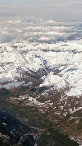 Vertical Above the peaks with clouds, capped Alps with rugged mountain peaks extending across landscape. Higher elevations are covered in a light dusting of snow, giving mountains a frosted appearance