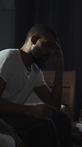 African American man expresses soul pain suffering from depression in dark bedroom. Guy with panic attack sits on unmade bed. Mental illness awareness