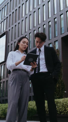 Busy Asian employees examine report on tablet standing by office building. Woman shows results to man partner discussing strategy. People reflect professionalism