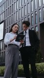 Busy Asian employees examine report on tablet standing by office building. Woman shows results to man partner discussing strategy. People reflect professionalism - Powered by Shutterstock - Get 15% off with code: PIKWIZARD15