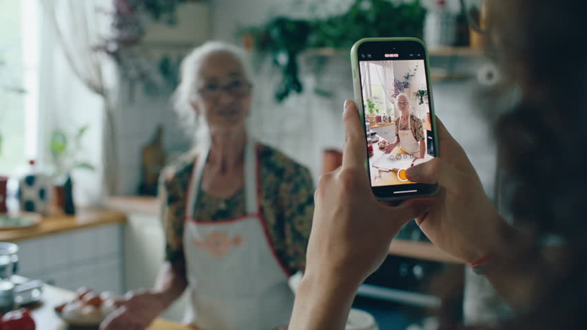 Close up view of boy holding smartphone and filming video of his elderly grandmother in apron explaining cooking recipe in kitchen