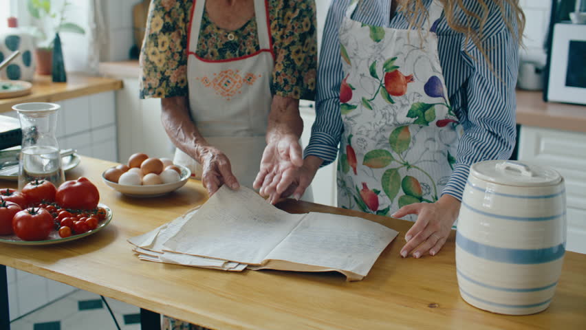 Senior grandmother in apron reading handwritten family recipe from book and explaining it to young granddaughter in cozy domestic kitchen