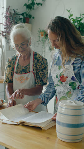 Elderly grandmother and her young granddaughter in apron looking through old handwritten recipe book, smiling and chatting as preparing to cook together in kitchen. Vertical clip