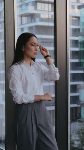 Young businesswoman stands by office window gazing thoughtfully at city skyline. Asian woman CEO contemplates future reflecting on opportunities and challenges