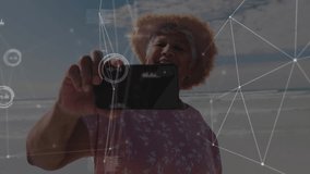 Senior woman lifting smartphone and showing technology on beach, with data overlay revealing view. Digital, wellness, lifestyle, vibrant, connectivity, leisure, modern - Powered by Shutterstock - Get 15% off with code: PIKWIZARD15