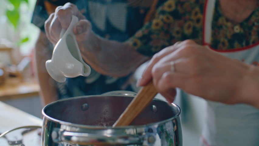 Hands of senior woman pouring olive oil into steaming pot as granddaughter stirring sauce with wooden spoon during home meal preparation in kitchen. Close-up view